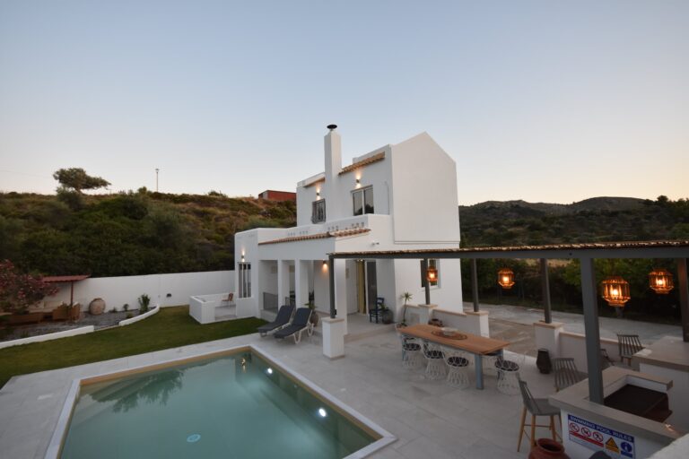 Evening view of Sofia Villa, a white villa with private swimming pool, outdoor dining pergola, and garden set against hillside scenery.